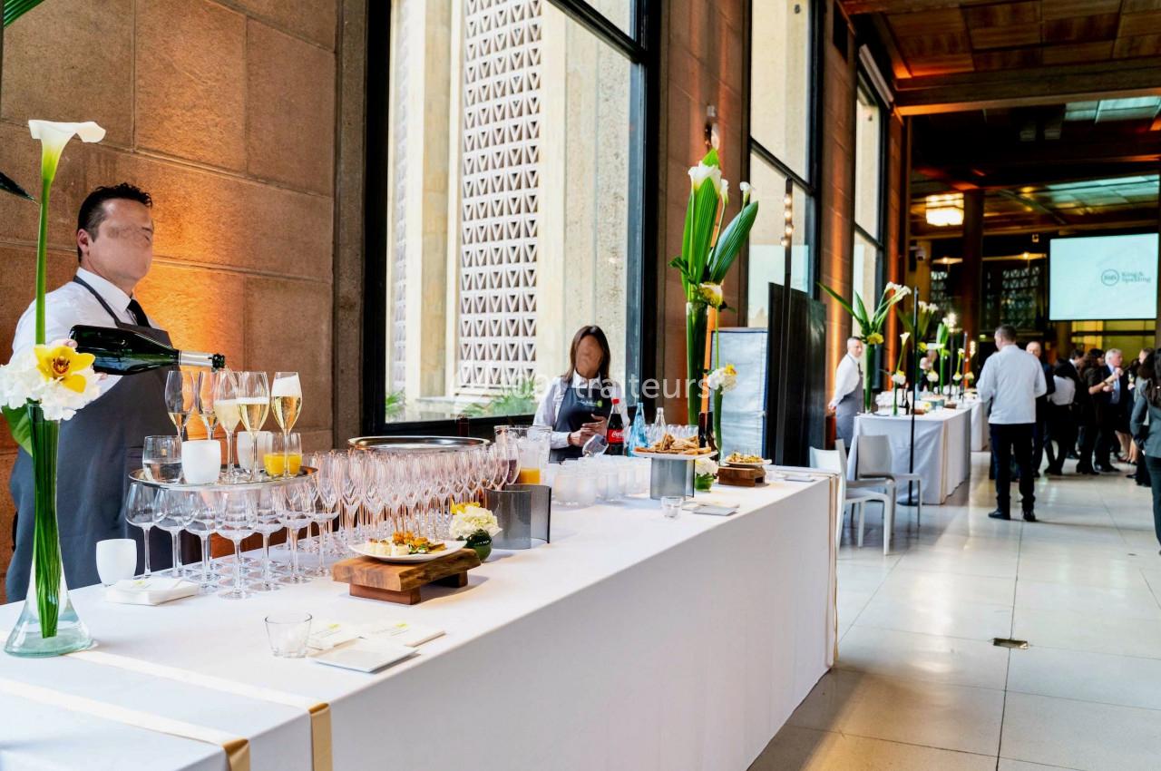 Buffet élégant avec boissons et amuse-bouches, décoré de fleurs, dans une salle lumineuse avec des invités en arrière-plan.