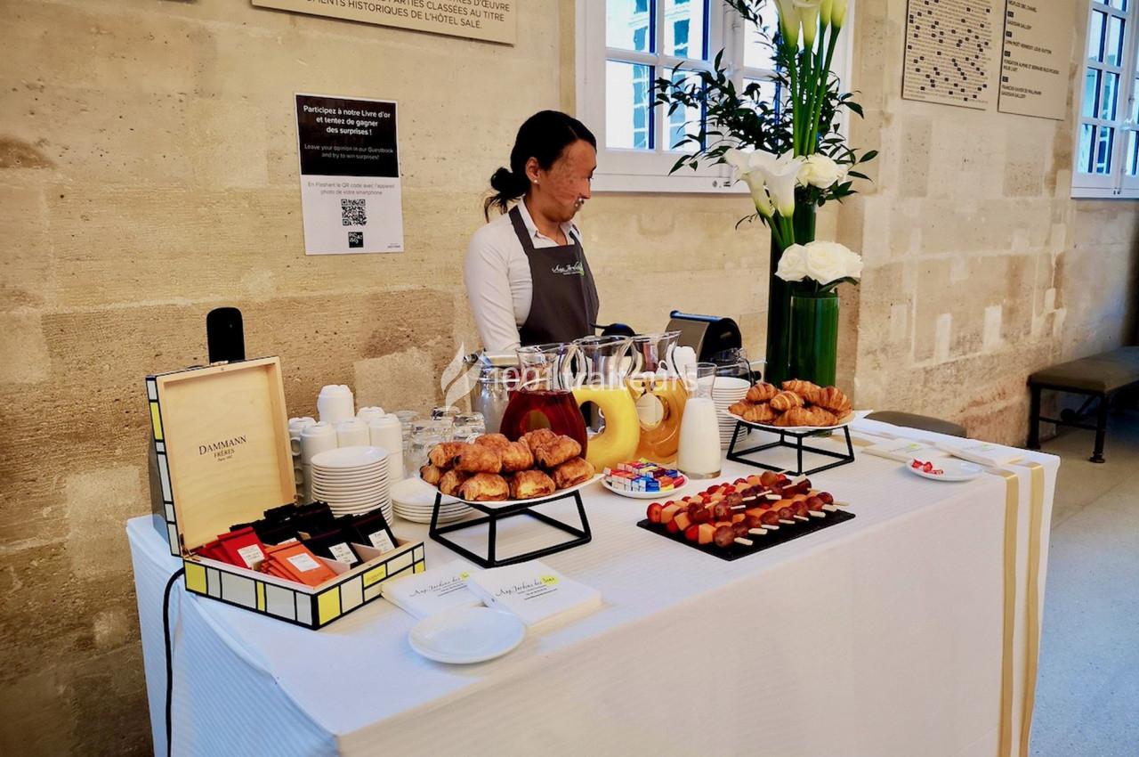 Table de buffet avec viennoiseries, boissons chaudes, jus de fruits et desserts, servie par une personne dans un espace…