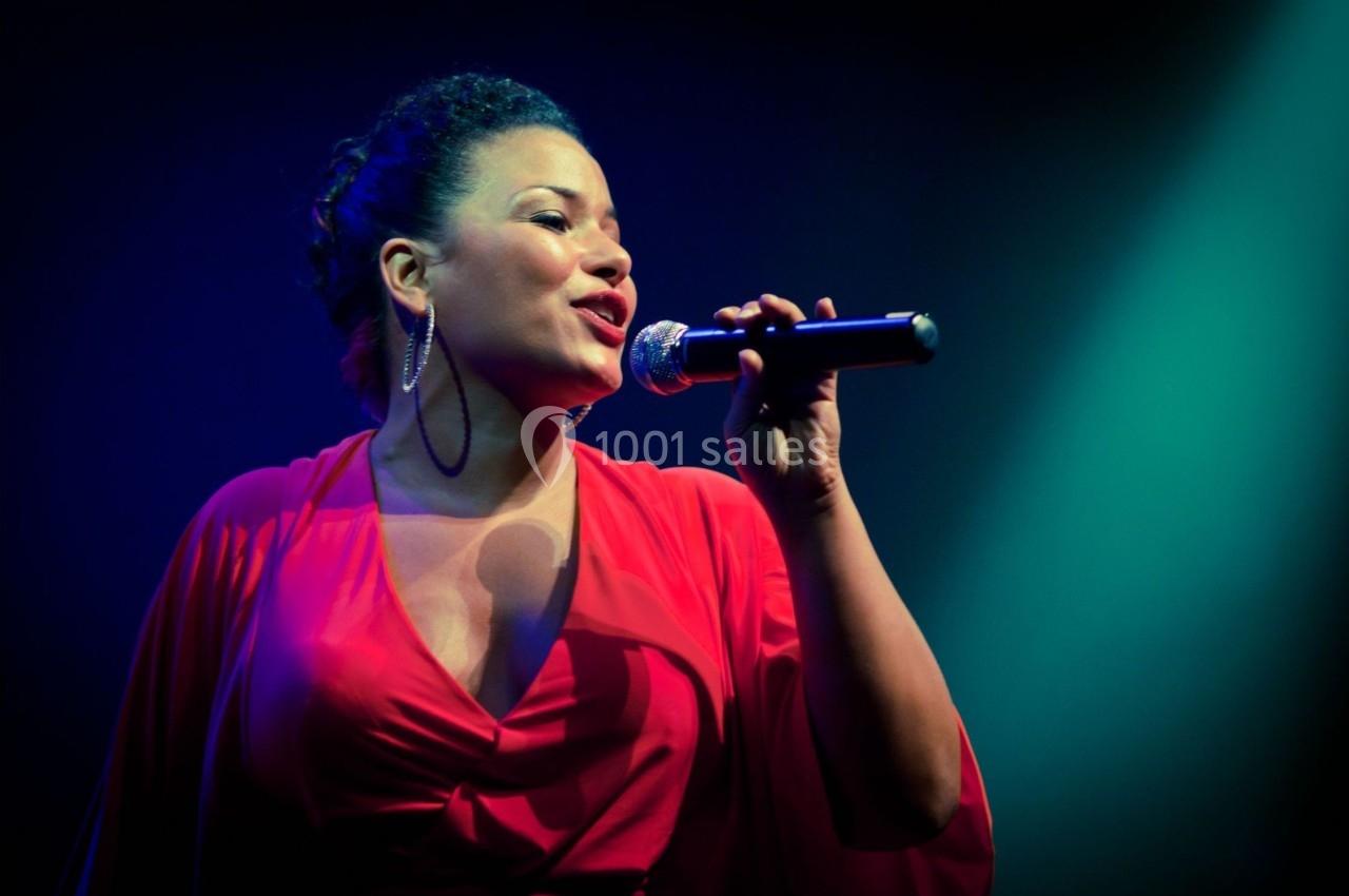 Femme en robe rouge chantant sur scène avec un micro, éclairée par des projecteurs colorés.