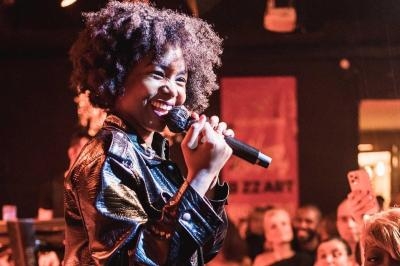 Une chanteuse souriante tenant un micro, sur scène devant un public attentif dans une ambiance chaleureuse.