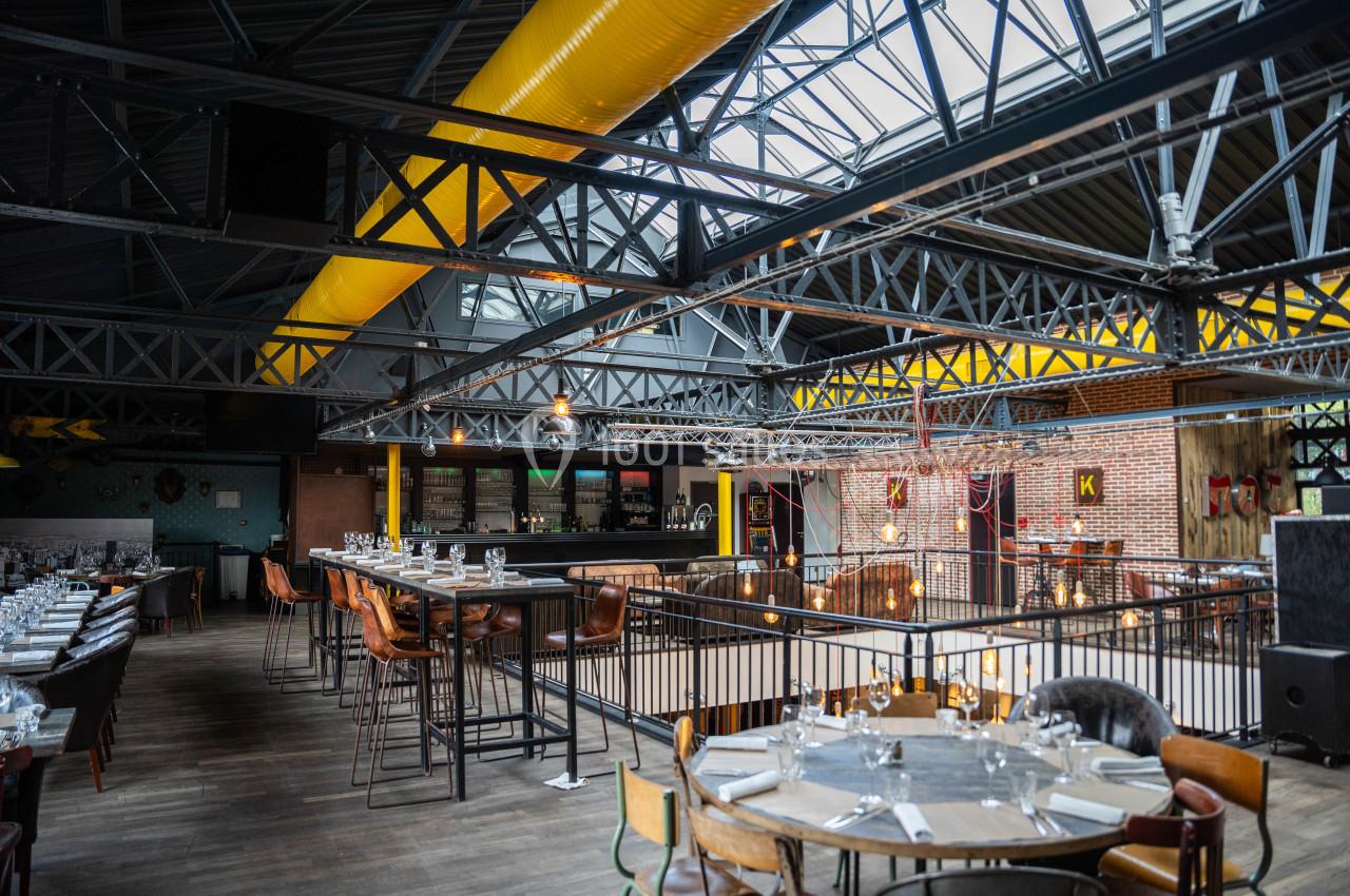 Intérieur d'un restaurant spacieux avec une structure métallique apparente, des tables dressées et un bar en arrière-plan.