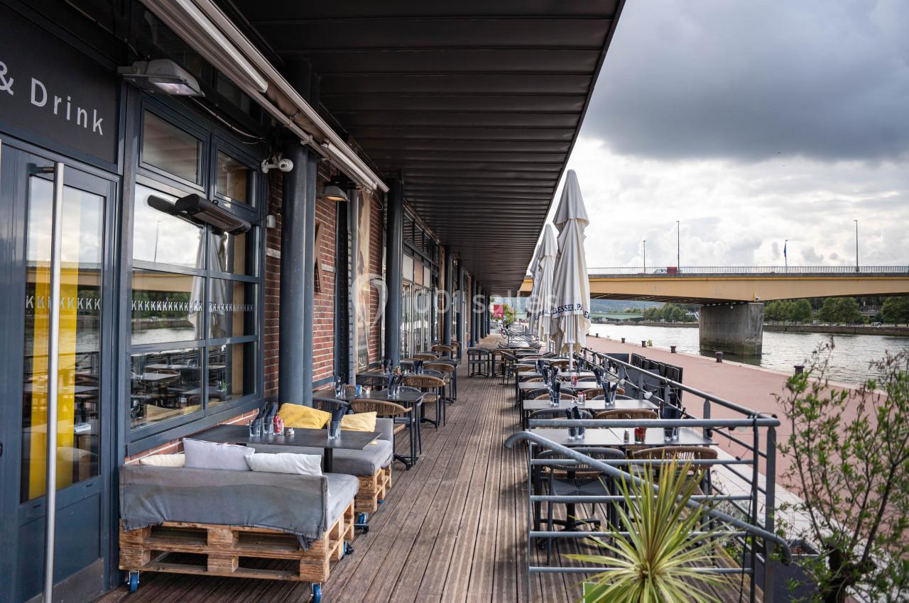 Terrasse en bois d'un restaurant au bord d'une rivière, avec tables, chaises et parasols sous un ciel nuageux.