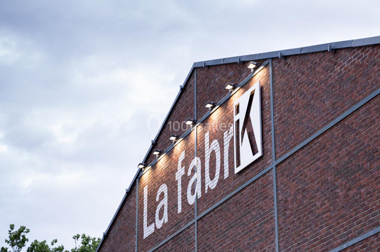 Façade en briques d'un bâtiment industriel avec l'inscription ’La Fabrik’ éclairée, sous un ciel nuageux.