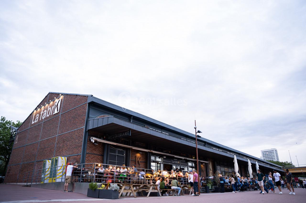 Façade d'un bâtiment en briques avec terrasse animée, des personnes assises et un ciel nuageux en arrière-plan.