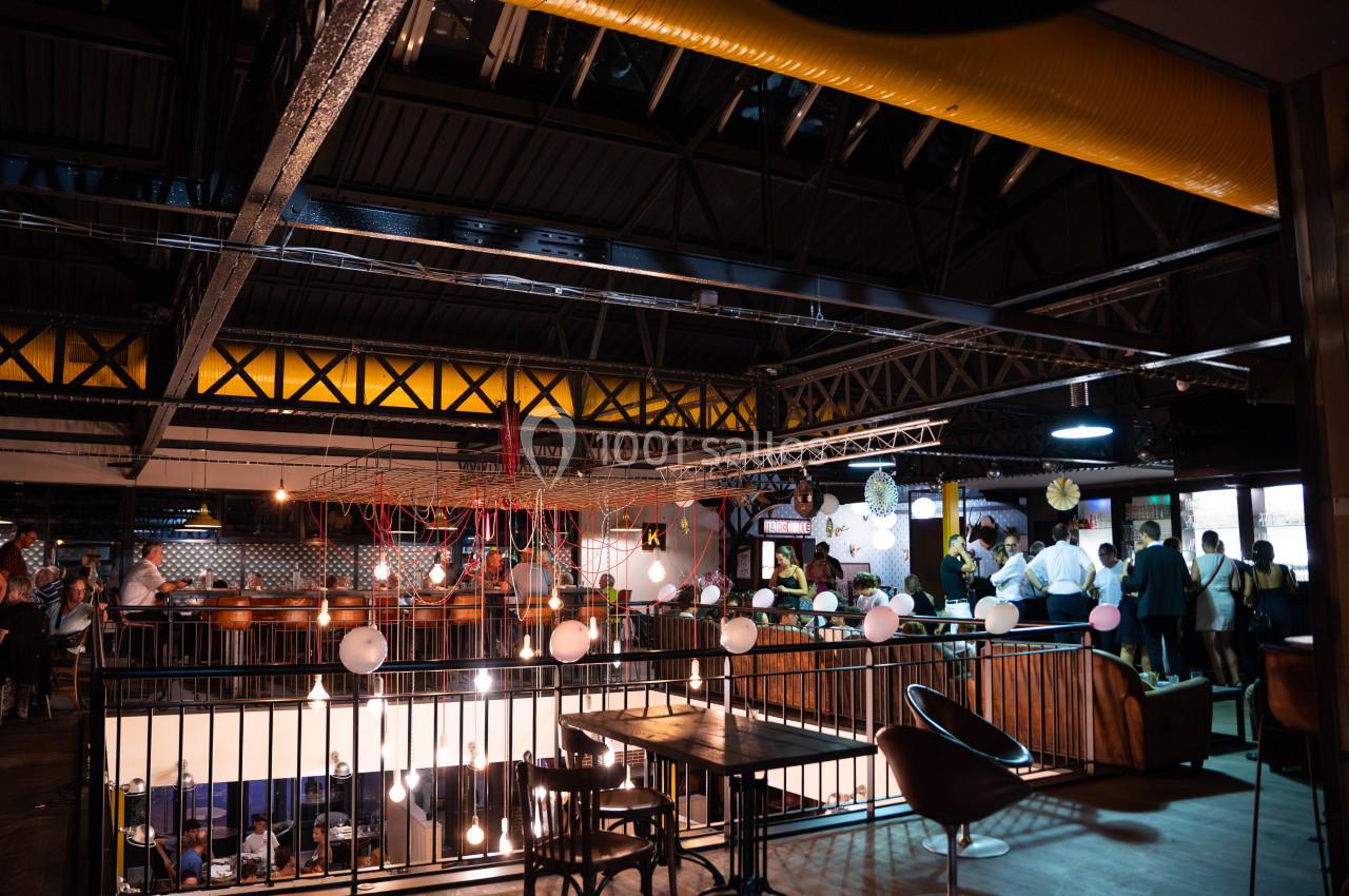 Espace intérieur industriel avec mezzanine, tables, chaises et éclairage tamisé, des personnes discutent en arrière-plan.