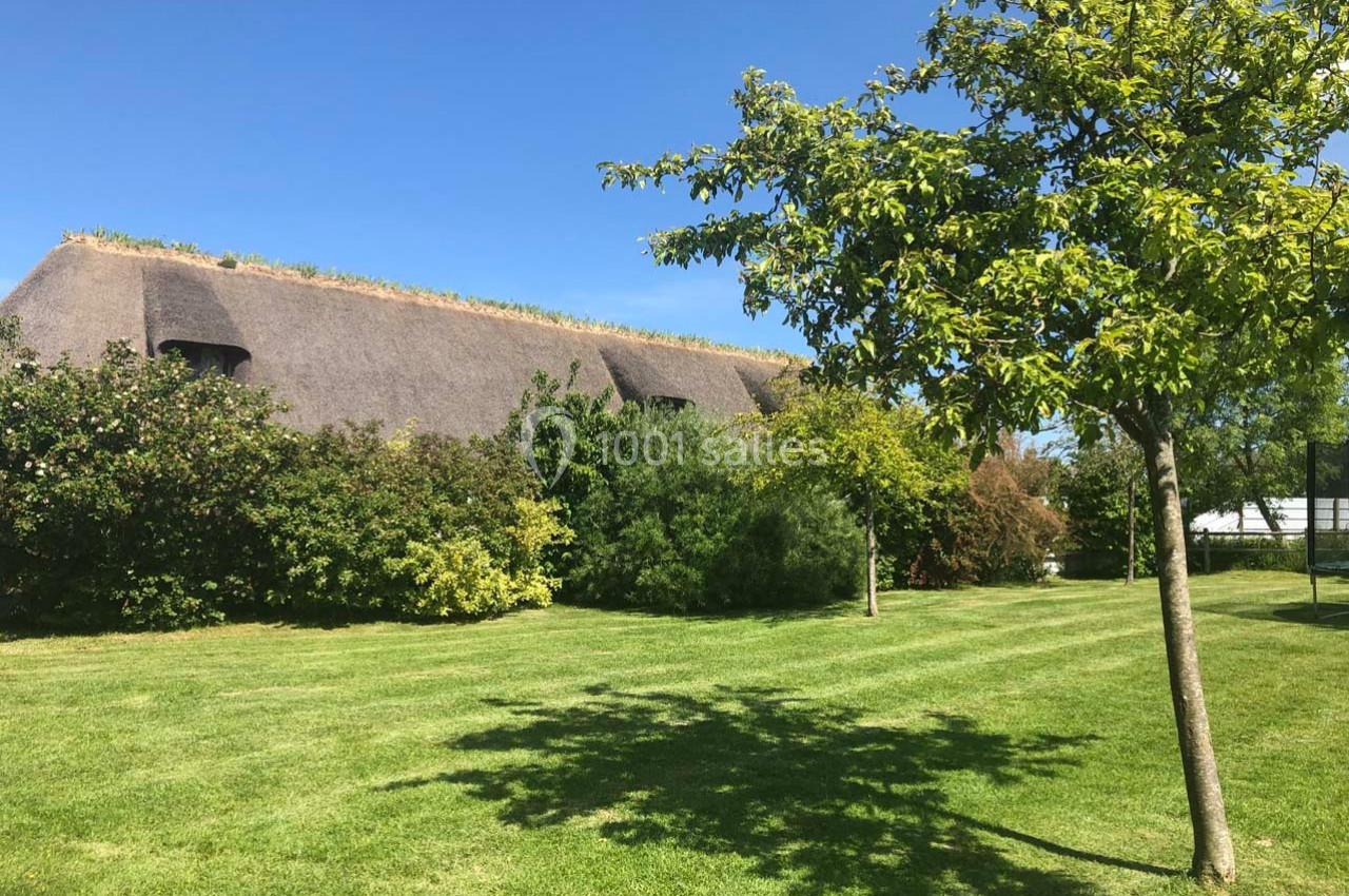 Toit de chaume d'une maison entourée d'arbres et d'arbustes, avec une pelouse bien entretenue sous un ciel bleu.