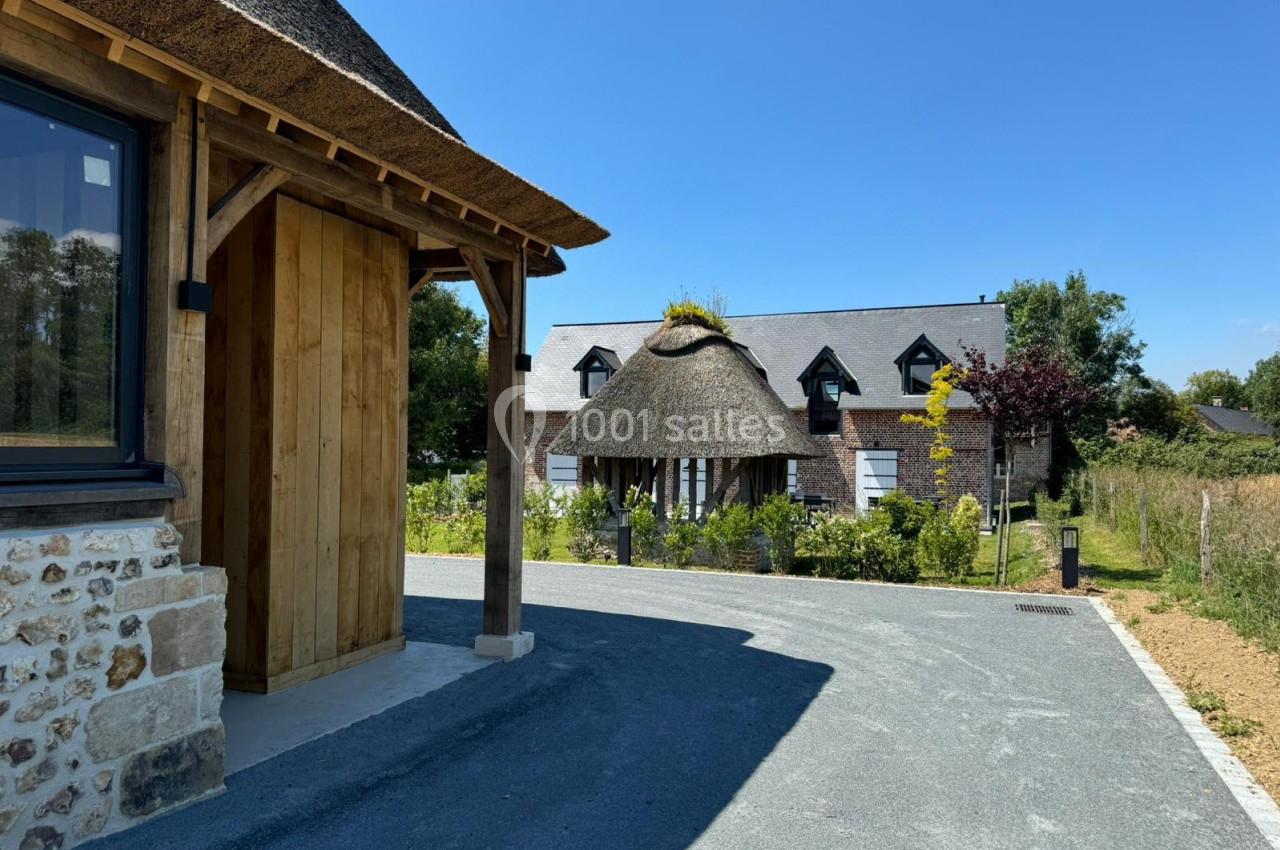 Maison en pierre et bois avec toit de chaume entourée de végétation, vue depuis une allée asphaltée.
