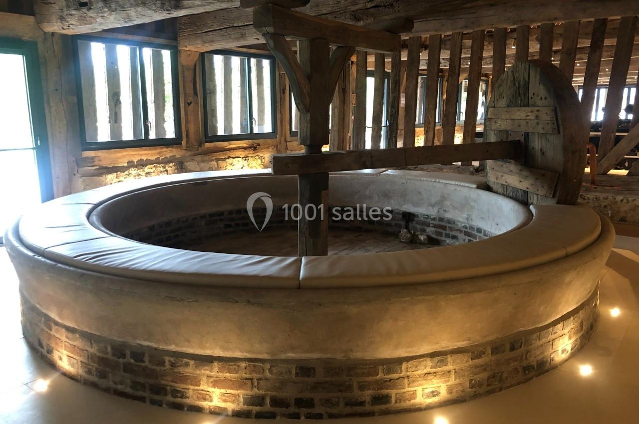 Pressoir ancien en bois et pierre dans une pièce lumineuse avec poutres apparentes et éclairage au sol.