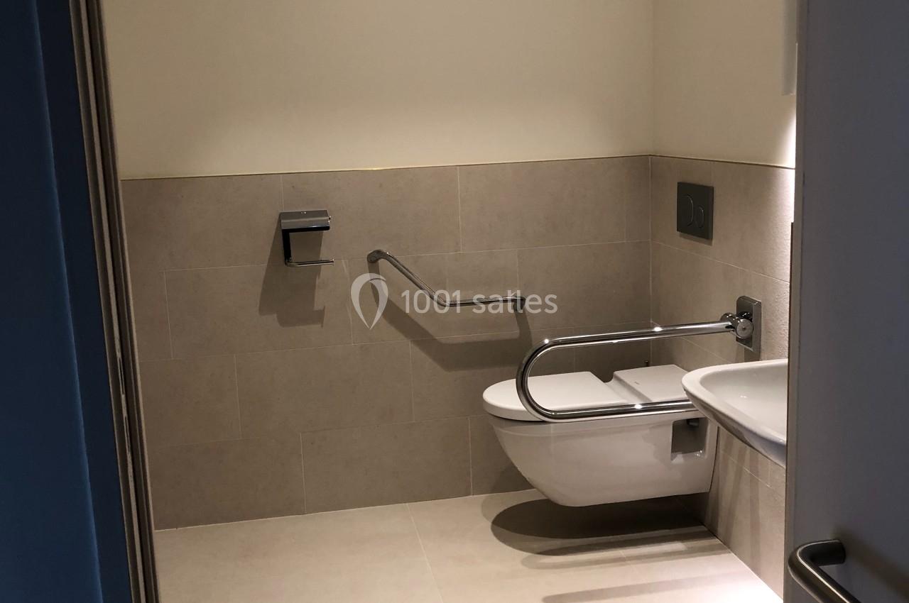 Toilettes accessibles avec barre d'appui, cuvette suspendue et distributeur de papier mural, éclairage sobre.