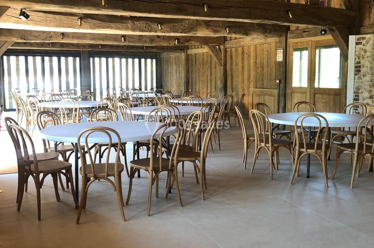 Salle avec charpente en bois apparente, tables et chaises en bois disposées dans un espace lumineux.