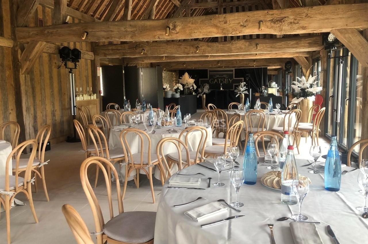 Salle de réception avec tables rondes dressées, chaises en bois clair et poutres apparentes dans un décor rustique.