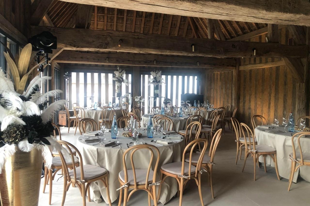 Salle de réception avec tables rondes dressées, chaises en bois courbé et poutres apparentes en bois.