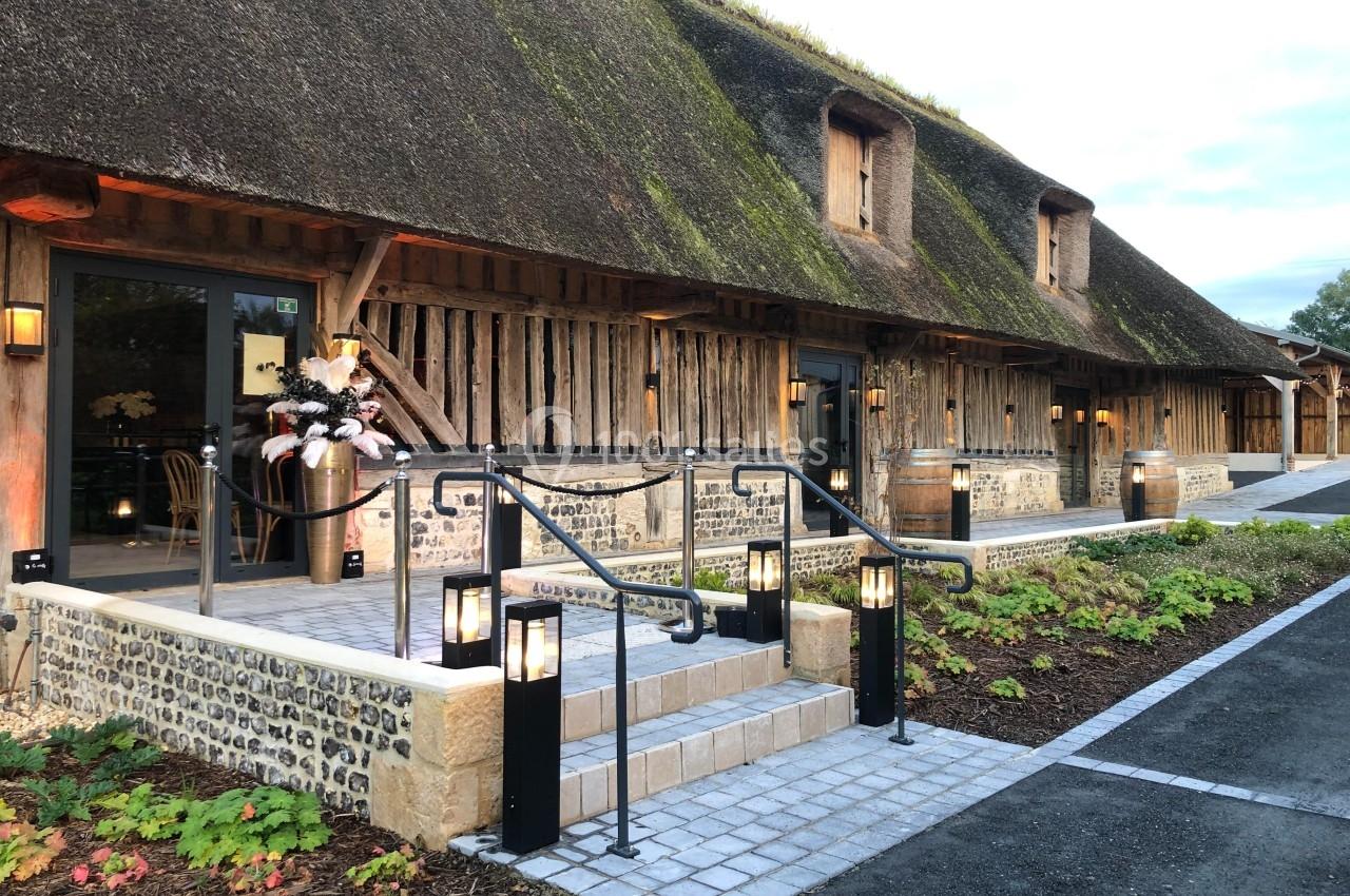 Bâtiment en colombages avec toit de chaume, éclairé par des lanternes, entouré de végétation et d'un chemin pavé.