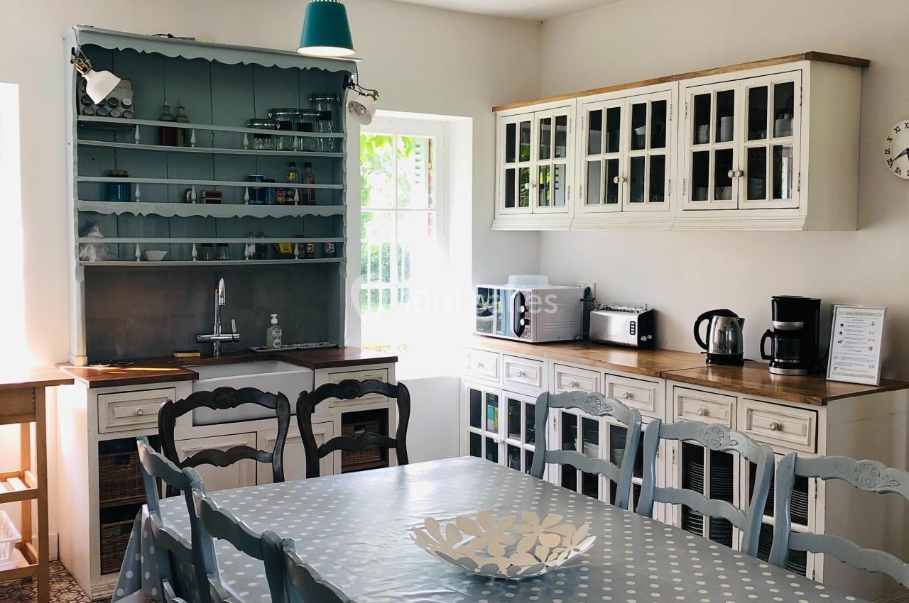 Cuisine lumineuse avec table en bois recouverte d'une nappe à pois, chaises bleues et meubles de rangement rustiques.