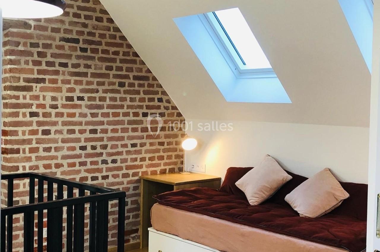 Chambre mansardée avec mur en briques, lit avec coussins, parquet clair et lumière naturelle provenant de deux fenêtres de…