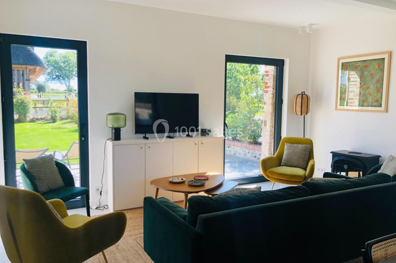 Salon lumineux avec canapé vert, fauteuils jaunes, table basse en bois et vue sur un jardin à travers de grandes baies…