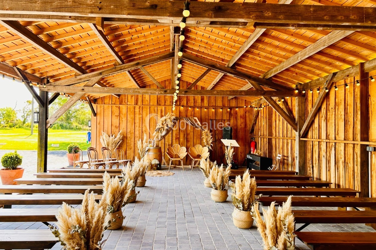 Salle de réception en bois avec bancs alignés, décorations en pampas et guirlandes lumineuses suspendues.