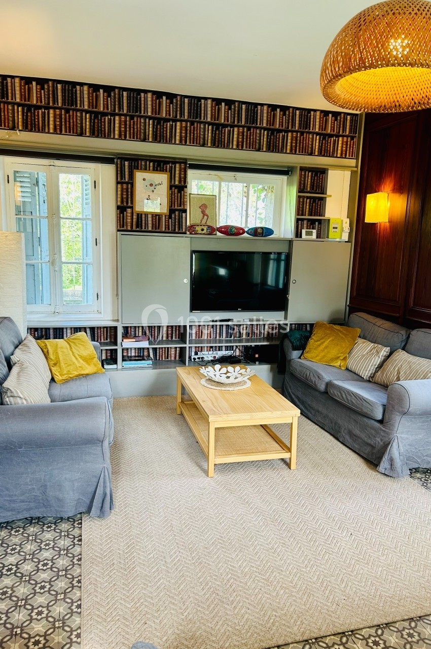 Salon avec deux canapés gris, table basse en bois, étagères remplies de livres et télévision murale.