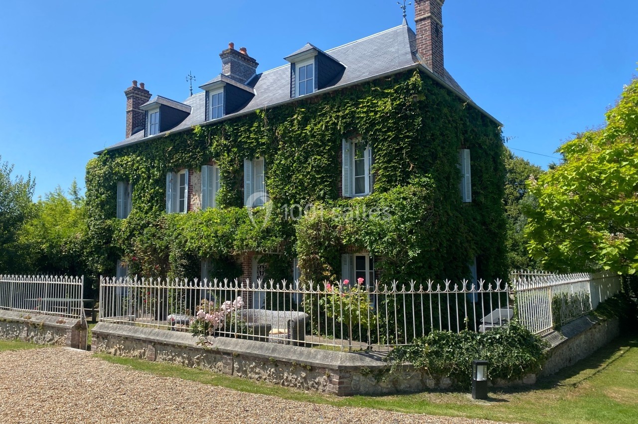 Maison ancienne en briques recouverte de lierre, entourée d'une clôture en fer forgé et d'un jardin fleuri.