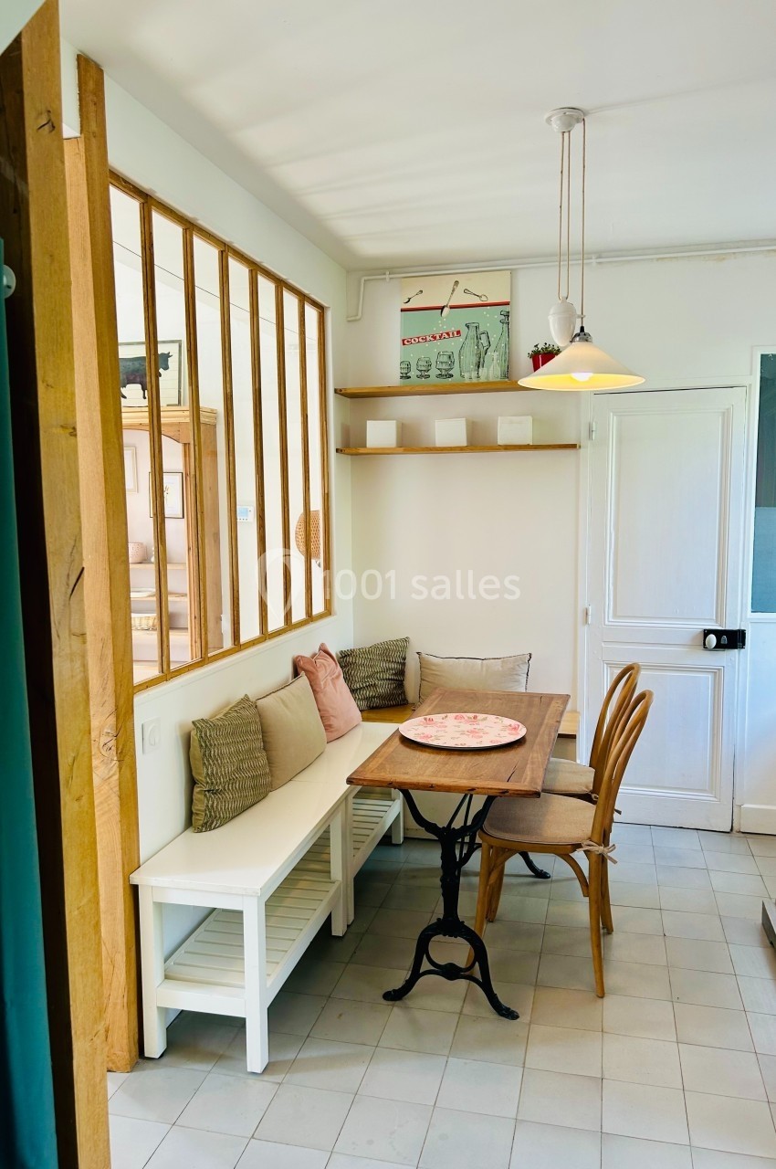 Coin repas lumineux avec table en bois, banc avec coussins, chaises, étagère murale et verrière intérieure.