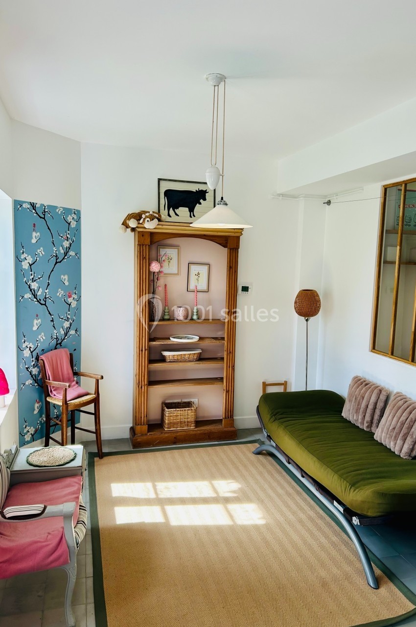 Salon lumineux avec canapé vert, étagère en bois, chaise rose, et mur décoré de papier peint fleuri.