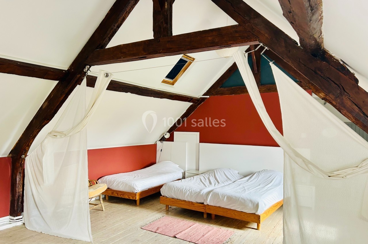 Chambre mansardée avec poutres apparentes, deux lits simples, murs rouges et blancs, sol en bois clair.
