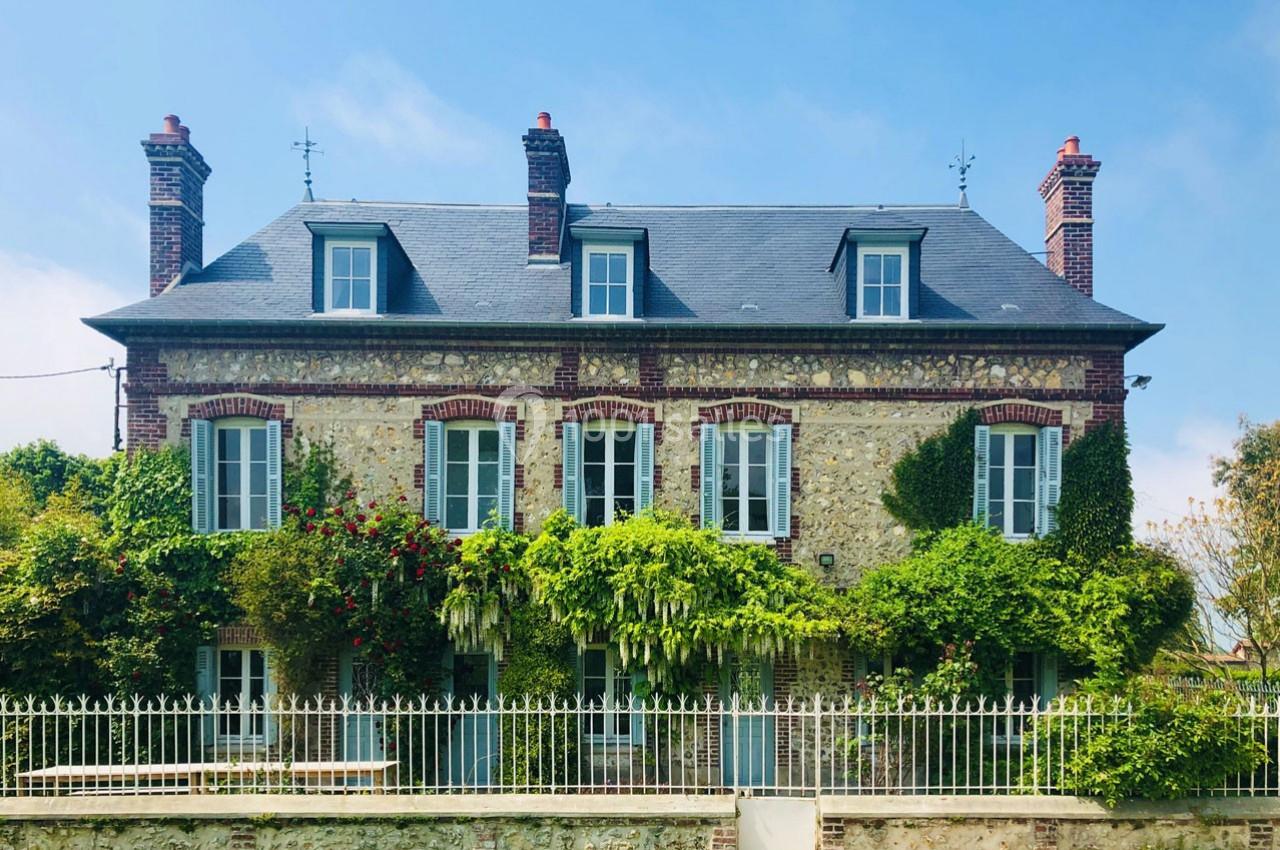 Façade d'une maison en pierre avec volets bleus, entourée de végétation et clôturée par une grille blanche.
