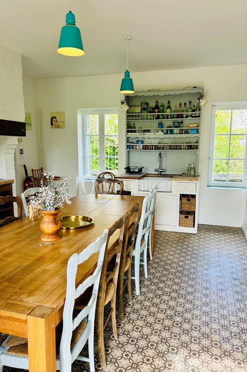 Cuisine lumineuse avec table en bois, chaises dépareillées, étagères ouvertes, et carrelage à motifs au sol.