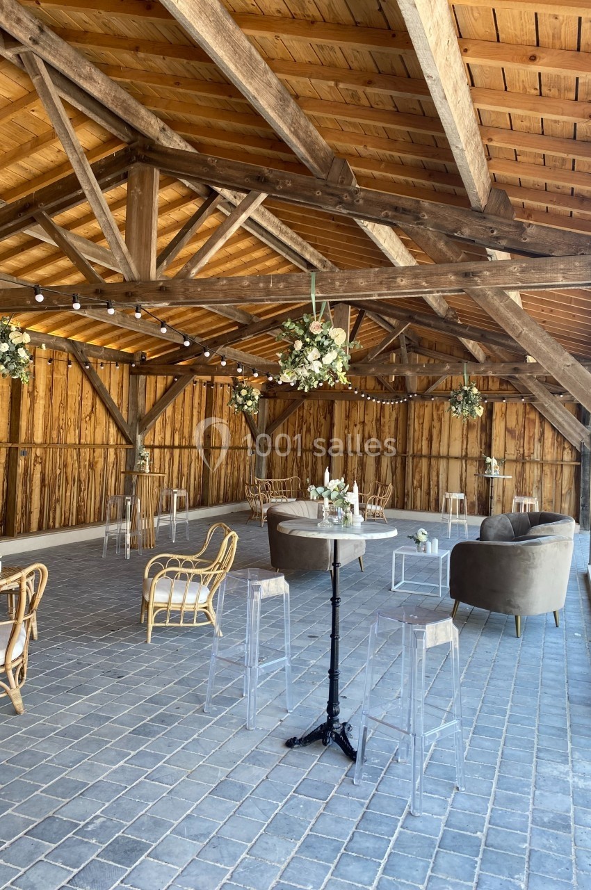 Salle en bois avec charpente apparente, aménagée avec des tables hautes, chaises en rotin et décorations florales suspendues.