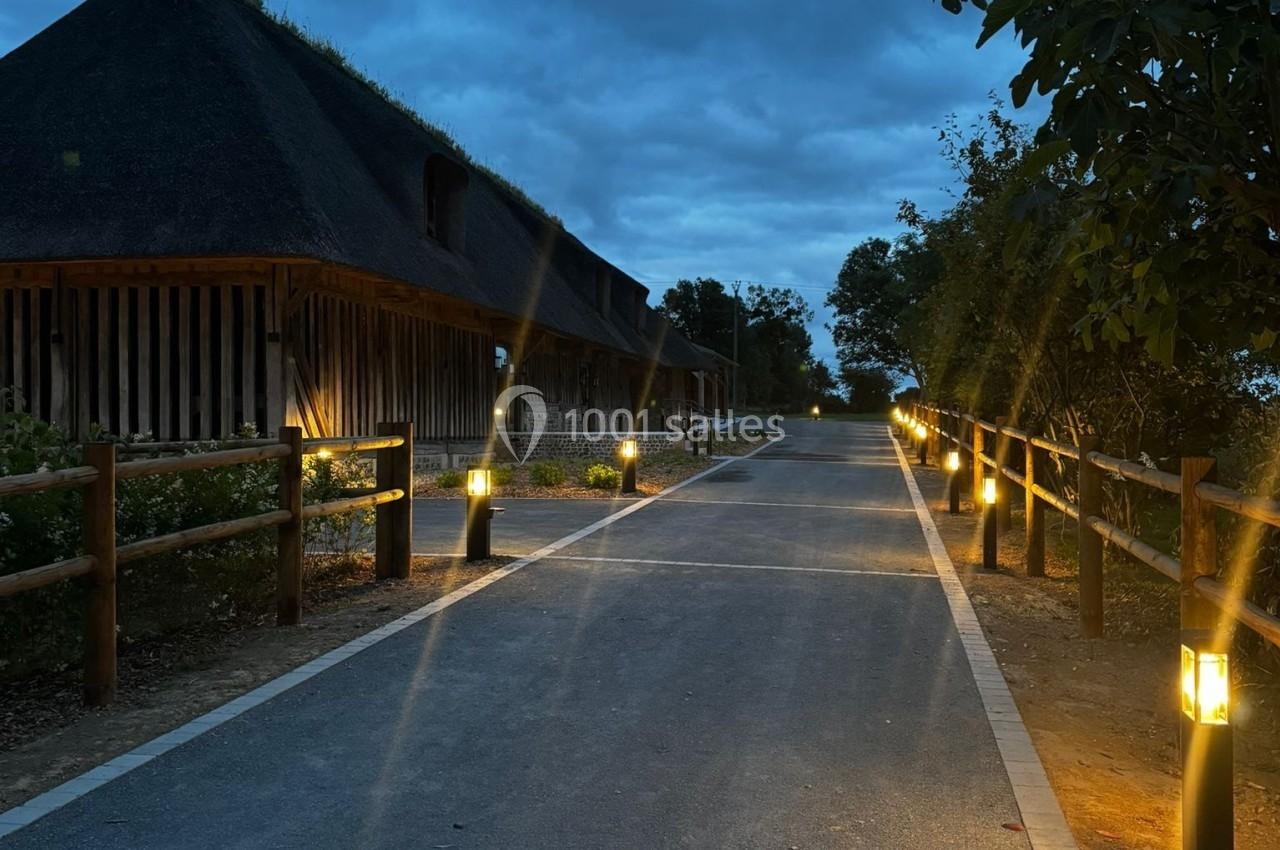 Chemin pavé bordé de lampadaires allumés, menant à des bâtiments en bois avec toits de chaume sous un ciel nuageux.