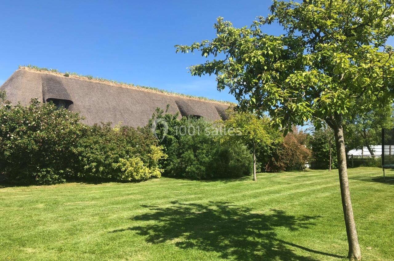 Toit de chaume entouré de végétation luxuriante, avec pelouse et arbre sous un ciel bleu clair.