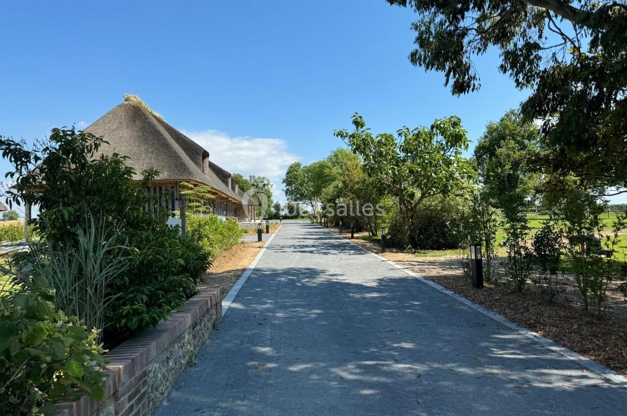 Chemin pavé bordé d'arbres et de végétation, avec un bâtiment au toit de chaume sur la gauche, sous un ciel bleu.