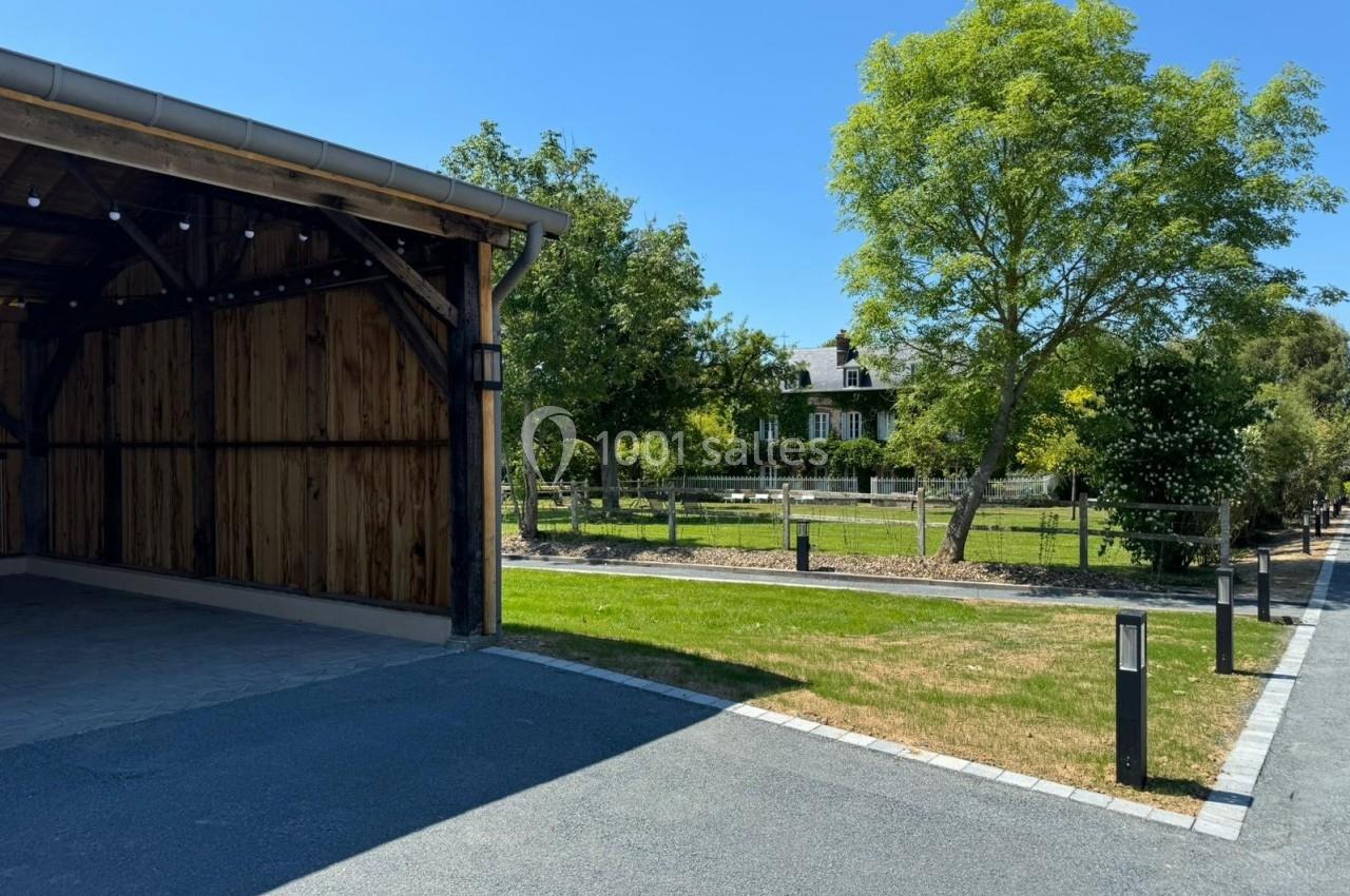 Abri en bois ouvert sur une allée pavée, avec pelouse, arbres et maison en arrière-plan sous un ciel dégagé.