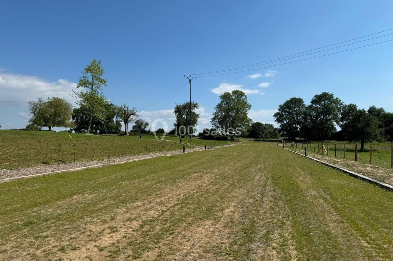Chemin herbeux bordé d'arbres et de clôtures, sous un ciel bleu avec quelques nuages.