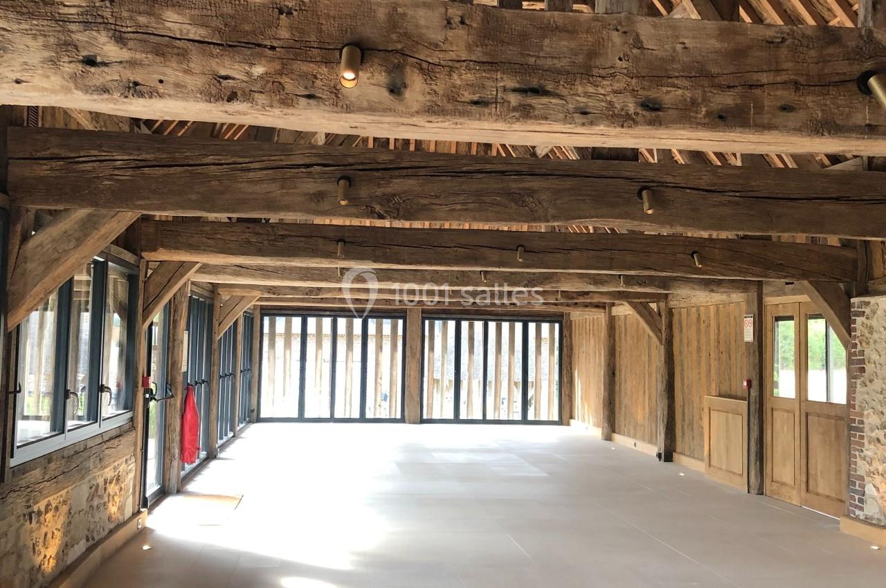 Intérieur d'une grande grange rénovée avec charpente en bois apparente et larges baies vitrées laissant entrer la lumière.