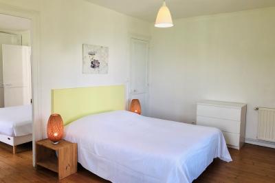 Chambre mansardée avec mur en briques, lit avec coussins, parquet clair et lumière naturelle provenant de deux fenêtres de…