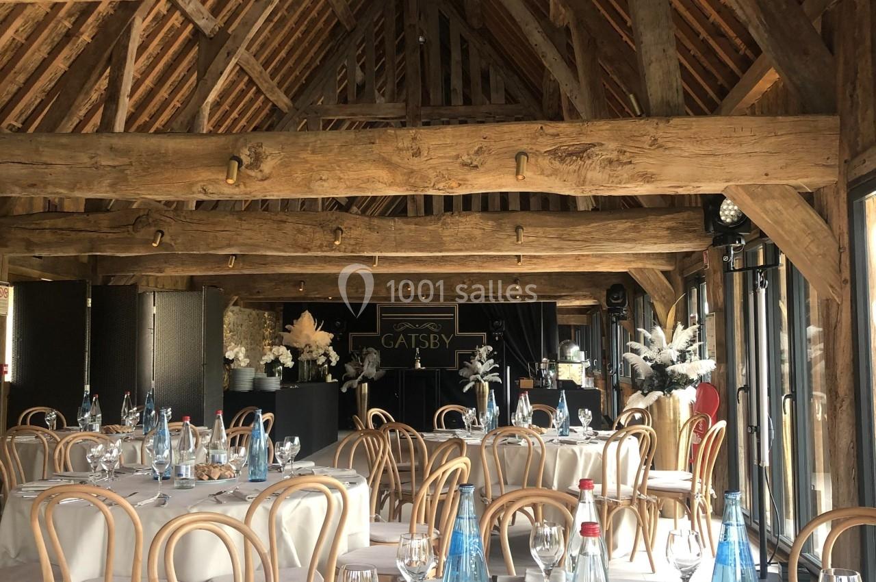 Salle de réception avec charpente en bois apparente, tables rondes dressées avec nappes blanches et bouteilles d'eau.
