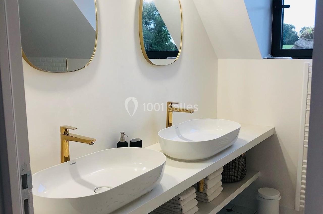 Salle de bain moderne avec deux vasques blanches, miroirs ovales dorés et éclairage mural sous un toit mansardé.