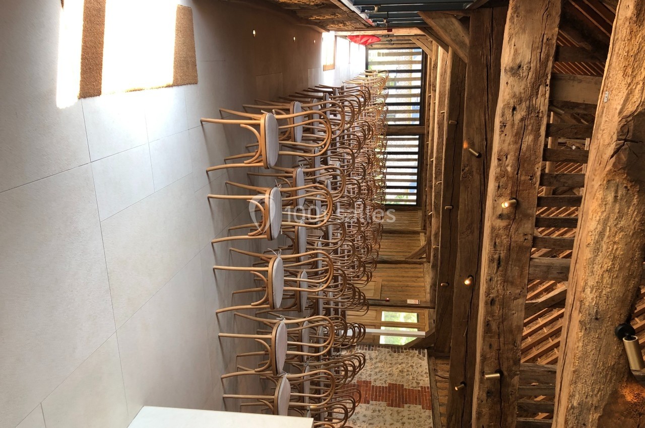 Salle avec des chaises empilées, poutres en bois apparentes et lumière naturelle entrant par de grandes fenêtres.