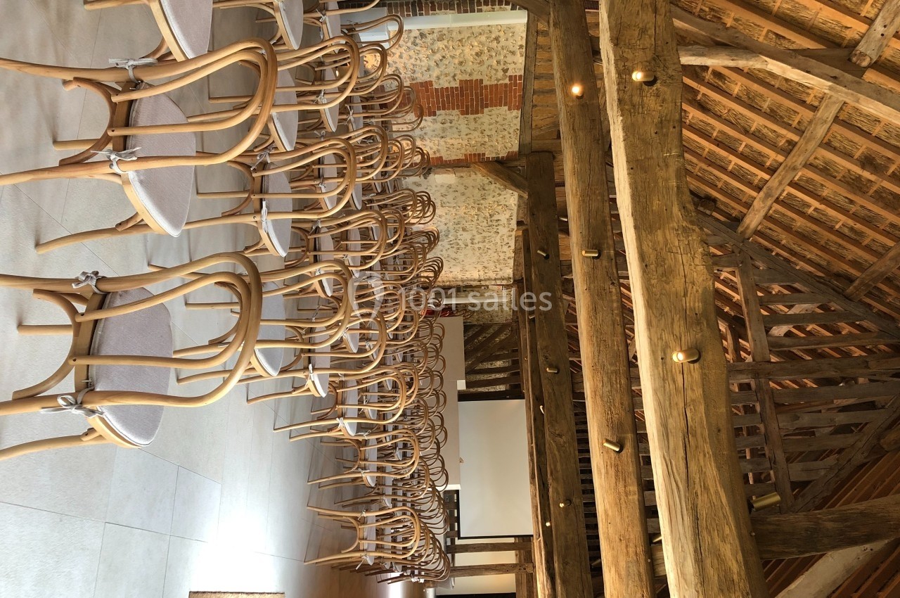 Rangées de chaises en bois clair alignées dans une salle avec poutres apparentes et murs en briques.