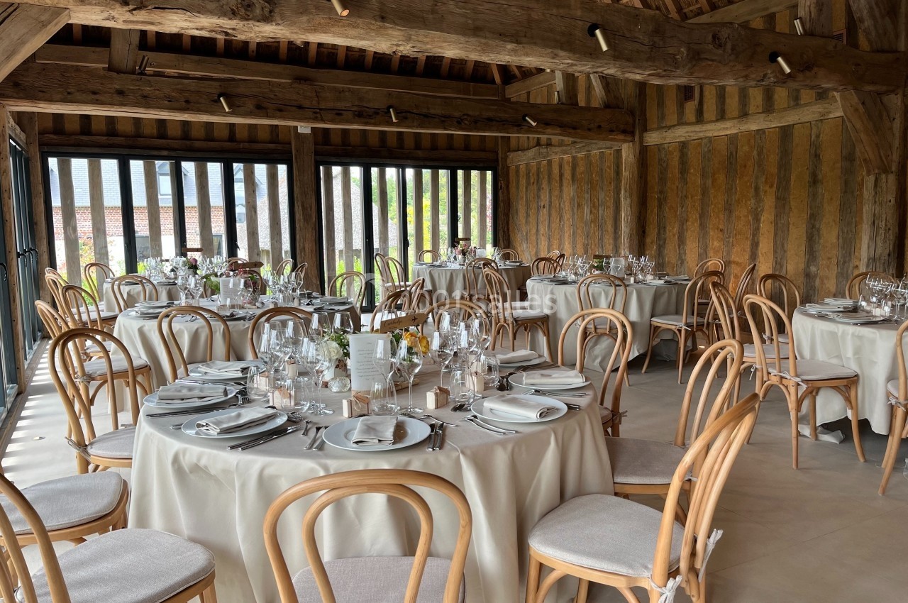 Salle de réception avec tables rondes dressées, nappes blanches, chaises en bois et grande baie vitrée donnant sur l…