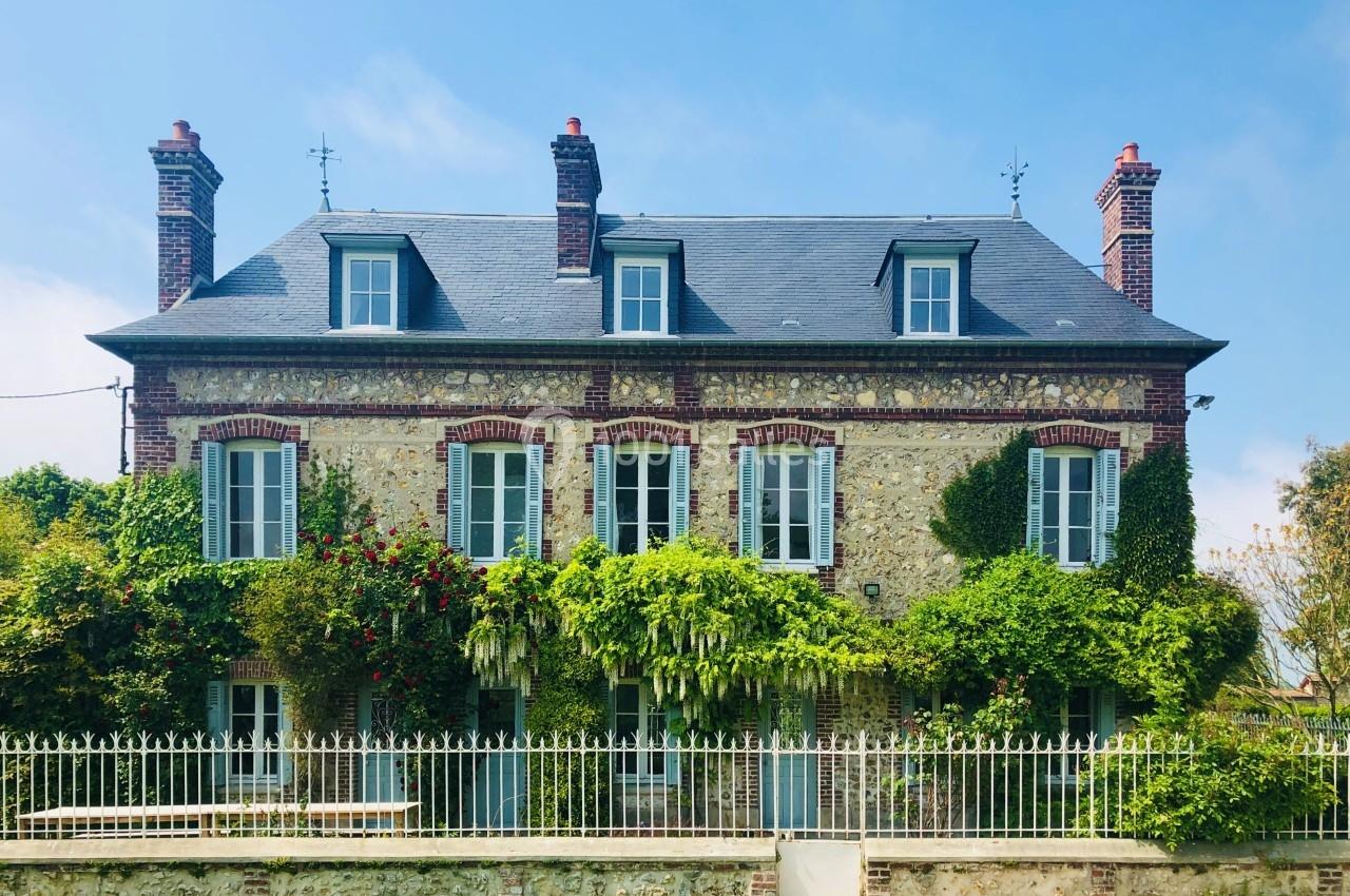 Façade d'une maison en pierre avec des volets bleus, entourée de végétation grimpante et clôturée par une grille blanche.