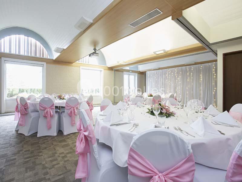 Salle de réception décorée avec des tables rondes, nappes blanches, chaises ornées de nœuds roses et rideaux lumineux.