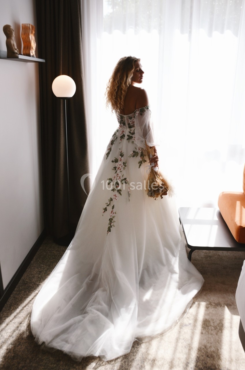 Femme en robe de mariée blanche à motifs floraux, tenant un bouquet, debout près d'une fenêtre lumineuse.