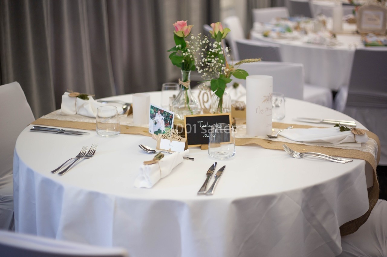 Table ronde décorée pour un événement, avec nappe blanche, fleurs, bougies et marque-place sur une bande en toile de jute.