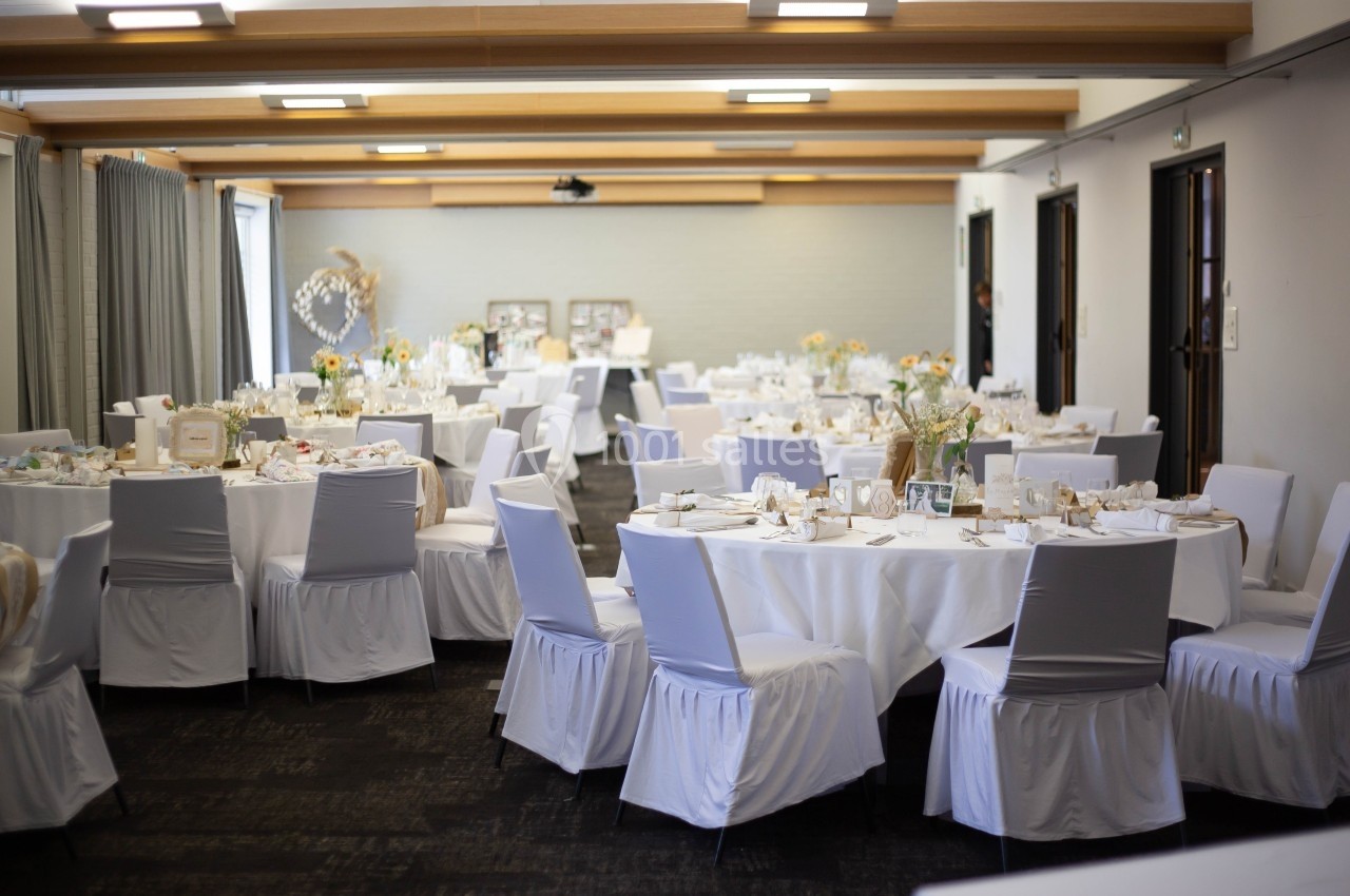 Salle de réception décorée avec des tables rondes, nappes blanches, chaises houssées et arrangements floraux.