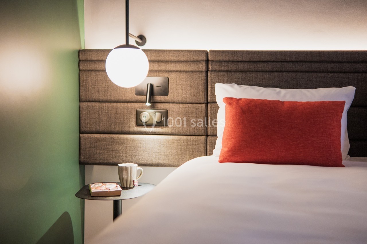 Lit avec tête de lit rembourrée, coussin rouge, lampe suspendue et table de chevet avec tasse et livre.