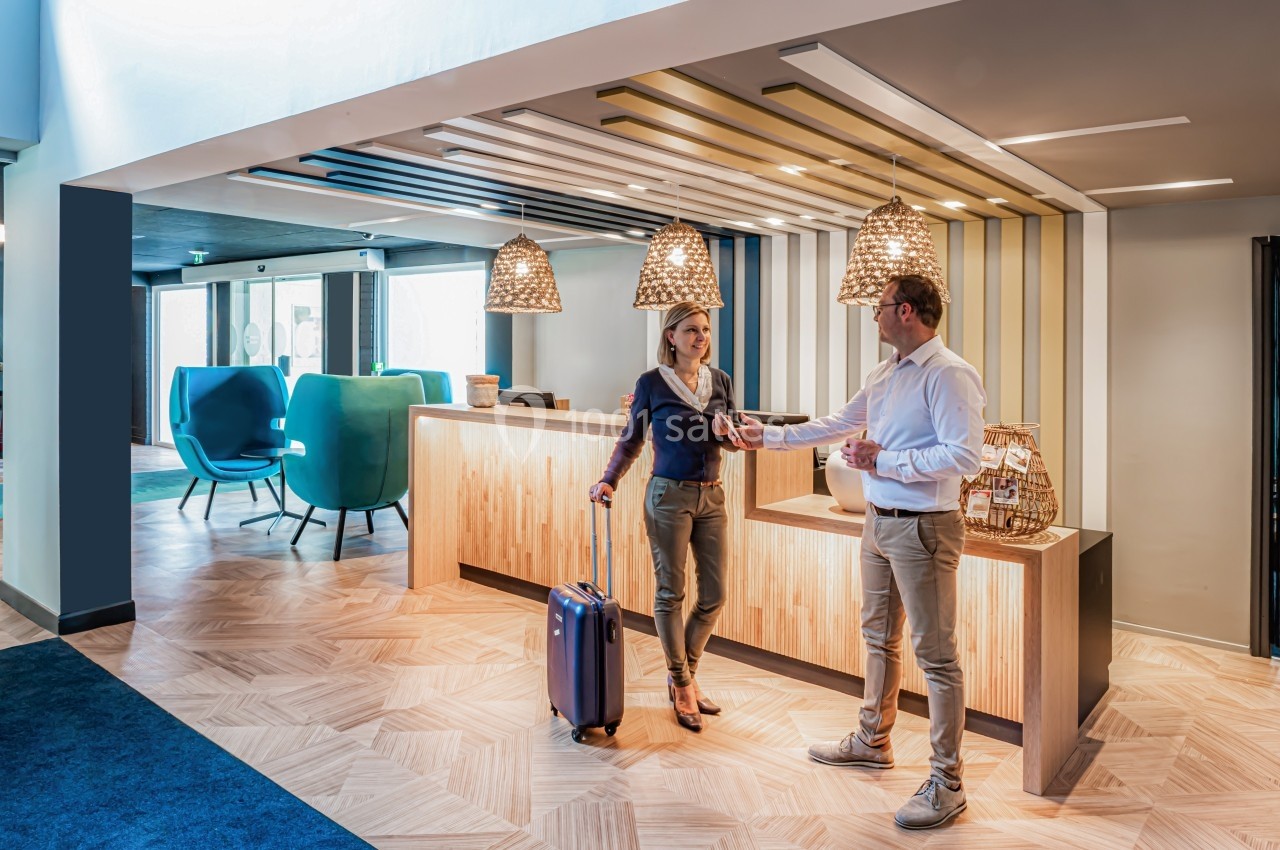 Un homme accueille une femme avec une valise dans un hall d'hôtel moderne et lumineux.