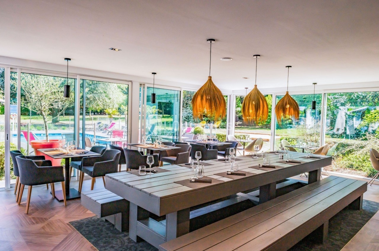 Salle lumineuse avec grandes baies vitrées, tables en bois, chaises modernes et vue sur un jardin avec piscine.