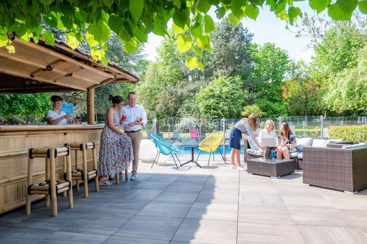 Personnes discutant et se détendant sur une terrasse près d'un bar en bois, avec piscine et végétation en arrière-plan.